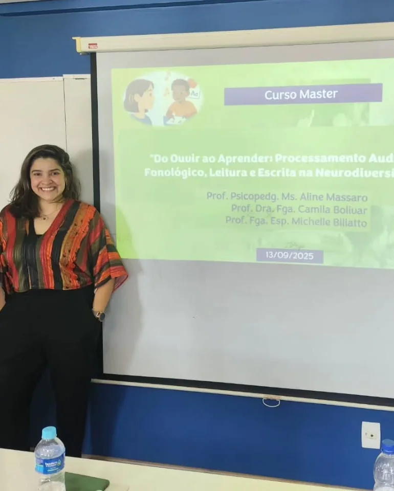 Dra. Camila Bolivar de pé, sorrindo, ao lado do telão com a abertura do Curso Master “Do Ouvir ao Aprender”, em 13 de setembro de 2025