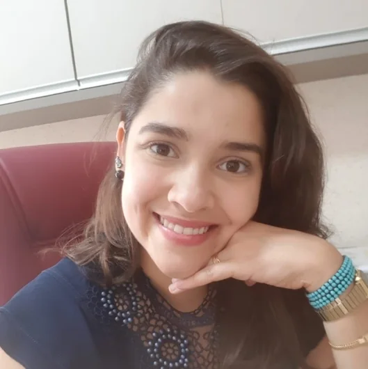 Dra. Camila Bolivar sorrindo, com a mão no queixo, sentada no consultório