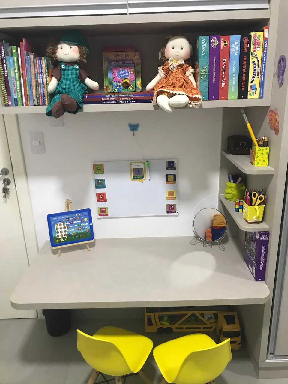 Cantinho com prateleira de livros infantis e bonecas de pano, quadro branco e duas cadeirinhas amarelas.