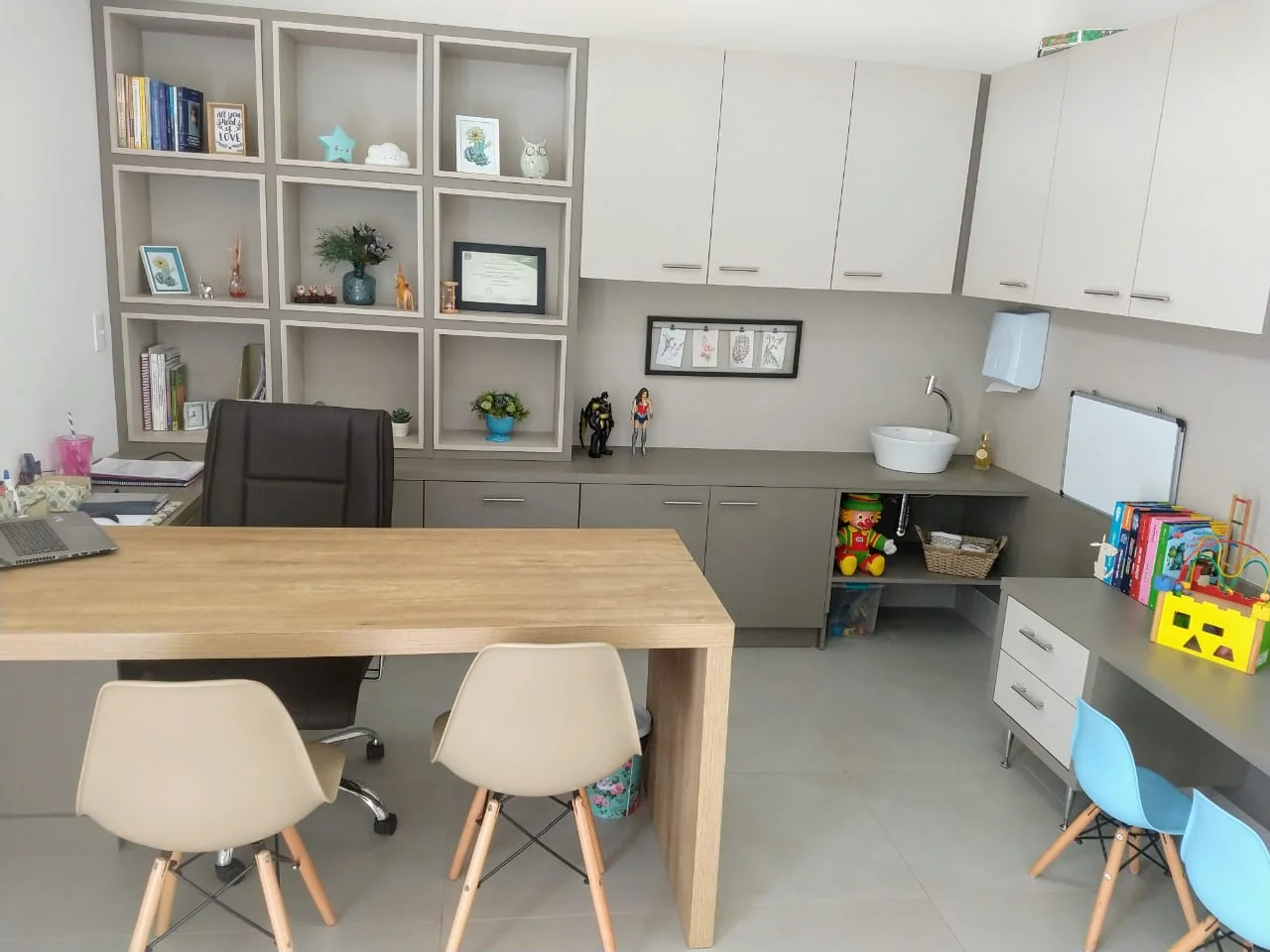 Consultório com mesa de madeira clara, estantes em nichos, pia e mesinha infantil com cadeiras azuis.