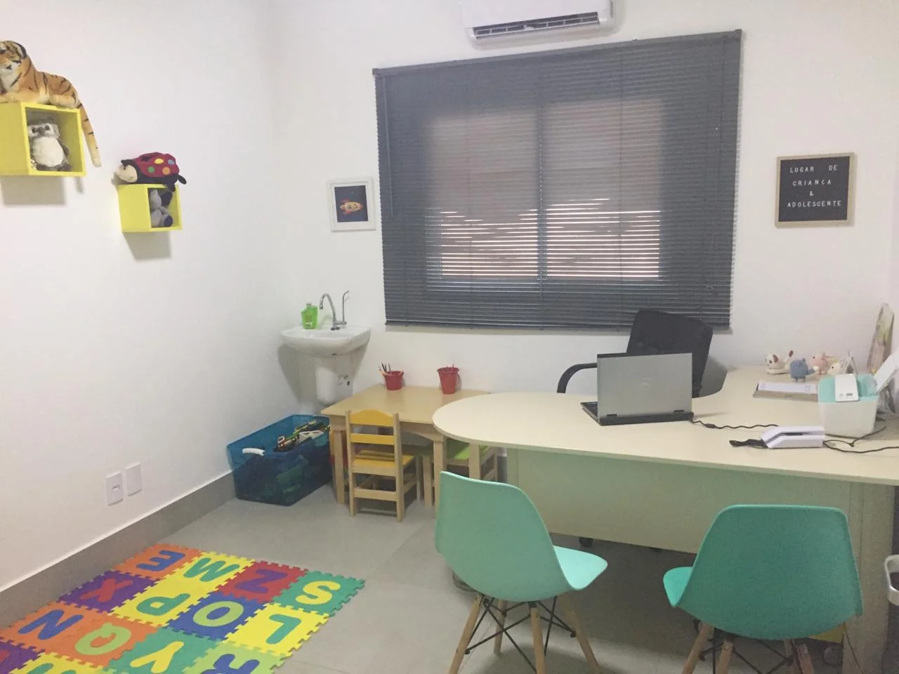Consultório claro da Incantare com tapete de letras em EVA colorido no chão, mesinha infantil de madeira, cadeiras verde-água e mesa de atendimento.