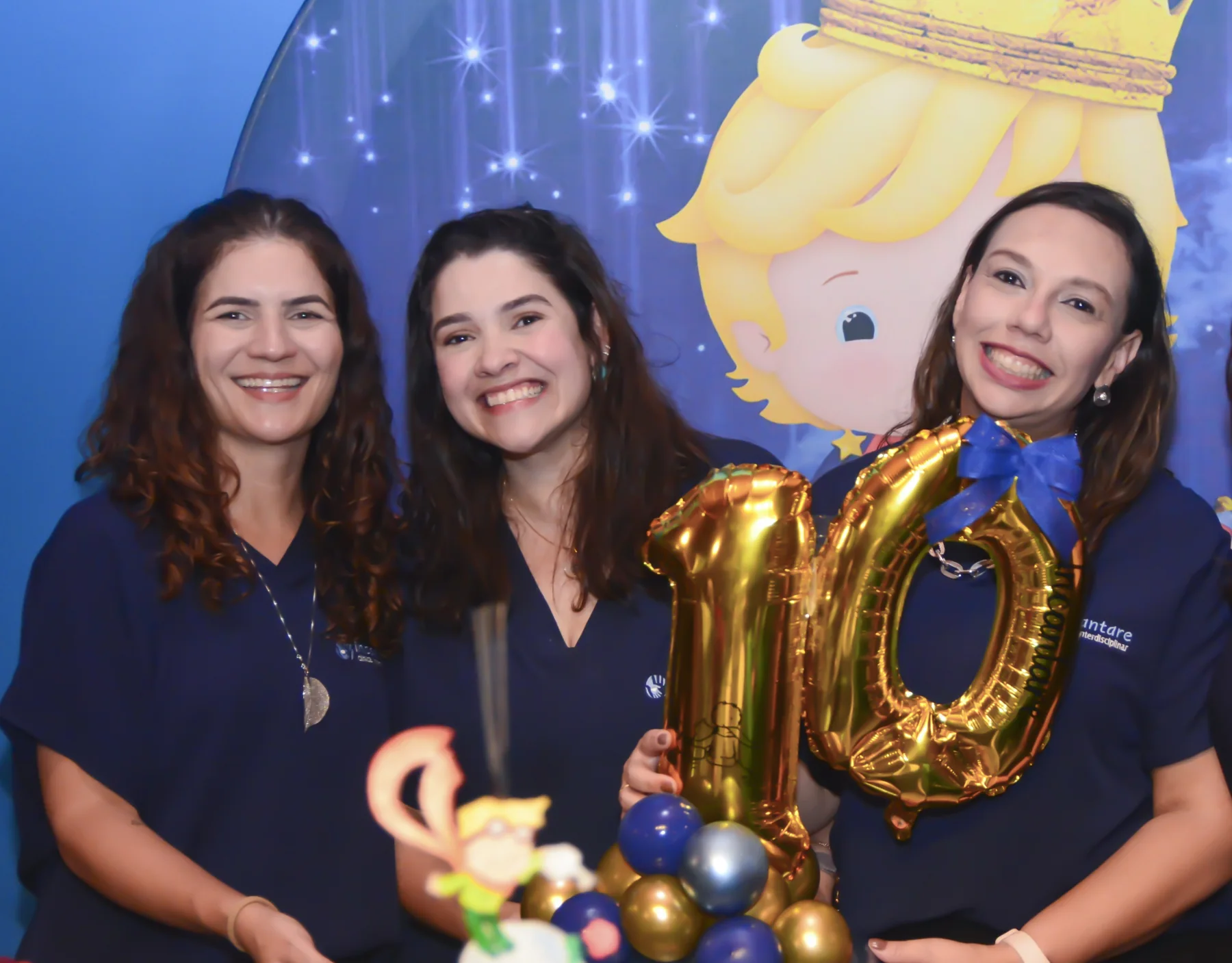 As três gestoras da Incantare, de uniforme azul-marinho, sorrindo diante de um painel azul estrelado na festa de 10 anos da clínica, com balões dourados do número 10; a Dra. Camila Bolivar está no centro.